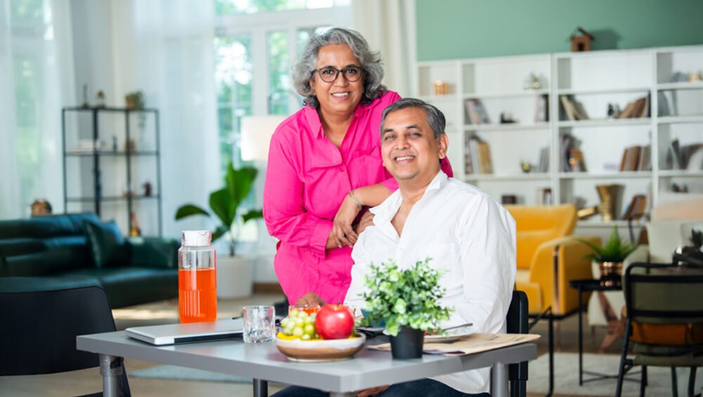 A smiling senior couple sitting together in a bright, modern living room, with fruits and a drink on the table, representing healthy and happy senior living.