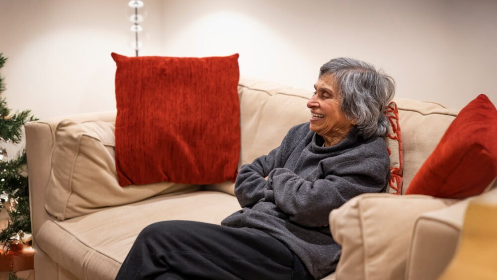 An elderly woman sitting comfortably on a sofa, smiling and relaxed in a warm, cozy living room setting.