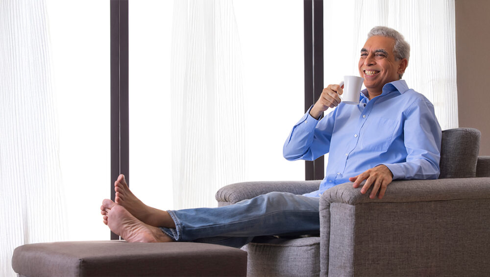 Smiling senior man relaxing on a chair with a cup of tea, representing comfort during a trial stay at Saral Satya Legacy senior citizen homes.
