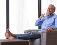 Smiling senior man relaxing on a chair with a cup of tea, representing comfort during a trial stay at Saral Satya Legacy senior citizen homes.
