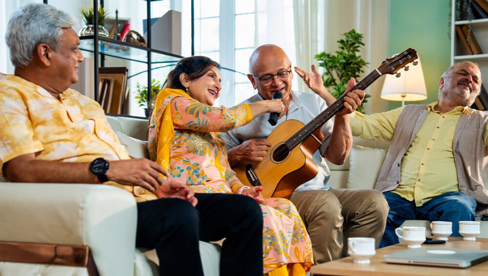 A group of seniors sitting together indoors, laughing and enjoying music as one plays a guitar and another sings, creating a joyful and lively atmosphere.
