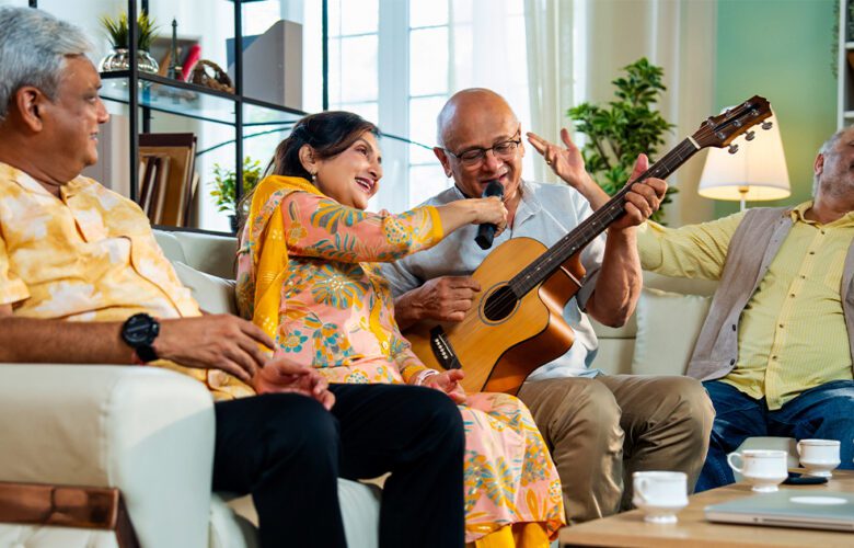 A group of seniors sitting together indoors, laughing and enjoying music as one plays a guitar and another sings, creating a joyful and lively atmosphere.