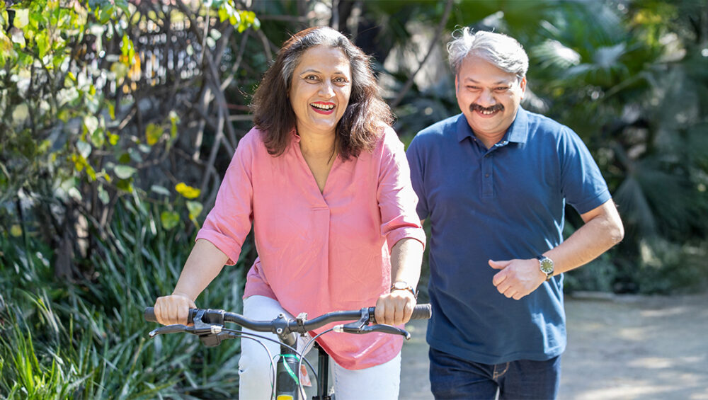 Smiling senior woman riding a bicycle with a companion walking beside her, showing active and independent living at Saral Satya Legacy.