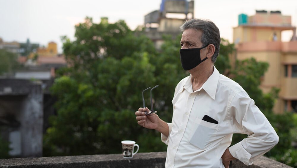 An older man wearing a mask standing outdoors on a terrace, holding his glasses and looking into the distance with buildings and trees in the background.