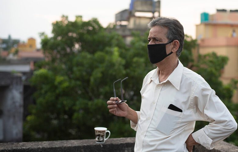 An older man wearing a mask standing outdoors on a terrace, holding his glasses and looking into the distance with buildings and trees in the background.