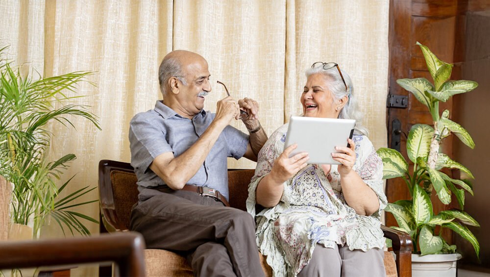 Smiling senior couple sitting indoors and using a digital tablet, reflecting comfort and safety provided by modern senior living homes at Saral Satya Legacy.