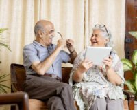 Smiling senior couple sitting indoors and using a digital tablet, reflecting comfort and safety provided by modern senior living homes at Saral Satya Legacy.