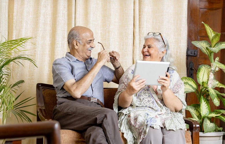 Smiling senior couple sitting indoors and using a digital tablet, reflecting comfort and safety provided by modern senior living homes at Saral Satya Legacy.