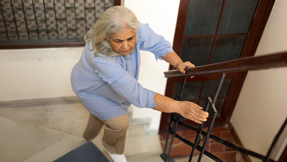 Senior woman carefully climbing stairs while holding a railing, representing the need to consider assisted living at Saral Satya Legacy.